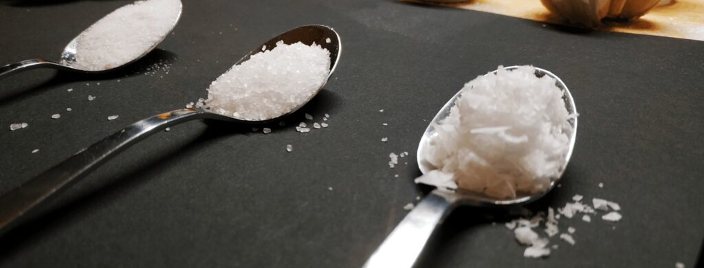 What is kosher salt? What is the difference between table salt and kosher salt? What is the difference between kosher salt and sea salt? This image features table salt, kosher salt and sea salt flakes in teaspoons
